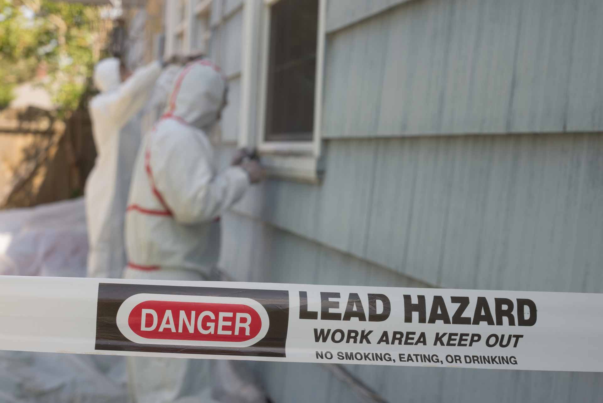 Workers in hazmat suit in a taped-off area. The tape reads, "Danger: Lead hazard. Work area. Keep out. No smoking, eating, or drinking."